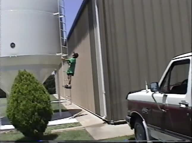 A young boy wearing a baseball cap fearlessly climbs the ladder of a tall grain silo on a farm