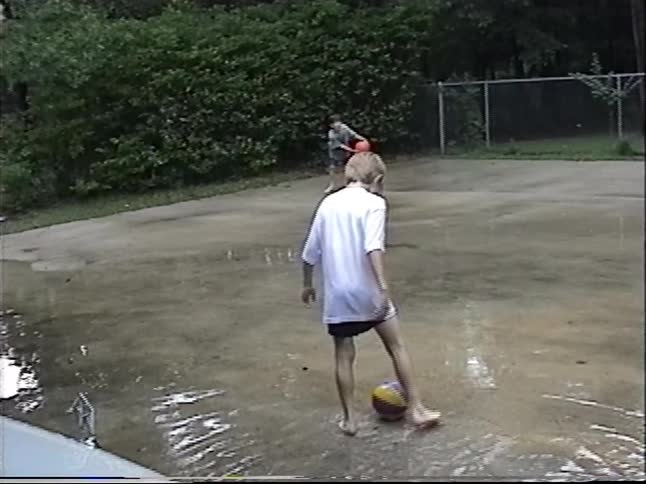 Two barefoot boys have fun playing basketball and splashing in puddles on their flooded driveway during a summer rainstorm