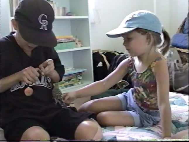 A young boy and girl sit on the floor of a bedroom and play with their pet parakeet in this vintage home video
