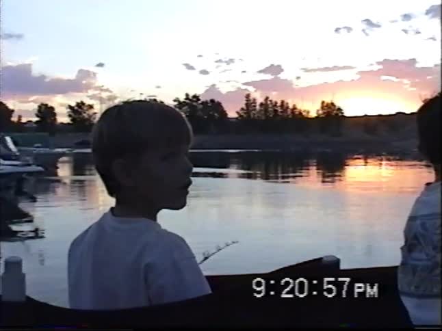 Two young boys enjoy an evening of fishing from a boat on a calm lake during a picturesque sunset