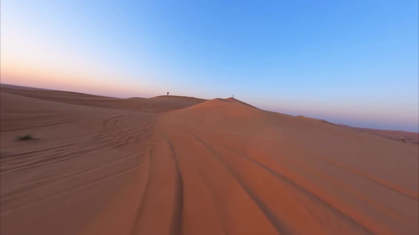 An aerial drone flies over the vast, rolling sand dunes of a desert at sunset towards people standing on a peak