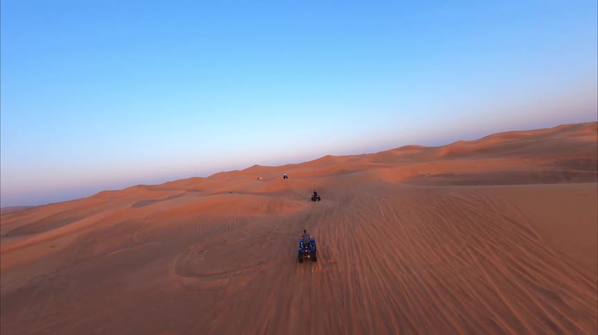 An exhilarating FPV follow shot of men riding ATVs in a line across the vast, golden desert sand dunes at sunset