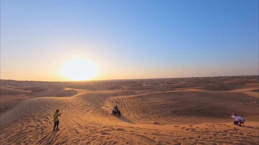 An aerial view follows a person riding a quad bike across expansive golden sand dunes as the sun sets