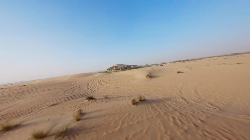 An aerial drone flies low across the vast, sandy desert toward a unique architectural structure under a clear blue sky
