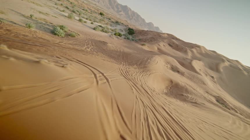 An immersive first-person view drone flight soars over vast, sun-drenched sand dunes covered in tire tracks, revealing a majestic desert mountain landscape