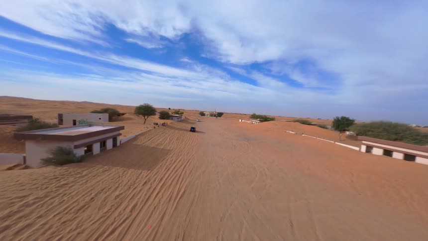 A dynamic FPV drone shot soars over an abandoned desert village, showing houses and a mosque being swallowed by the shifting sands