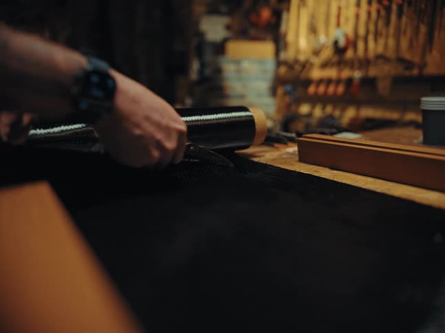 A close-up of a person's hands cutting a sheet of carbon fiber fabric with scissors in a workshop