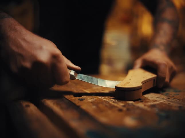 A craftsman with tattooed arms meticulously uses a carving knife to shape a piece of wood for a stringed instrument in their workshop