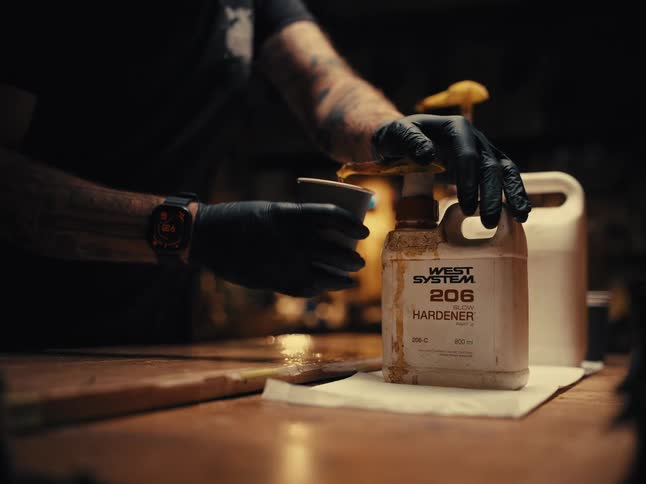 A tattooed craftsman wearing black gloves carefully pumps slow hardener into a cup on a wooden workbench in a dimly lit workshop