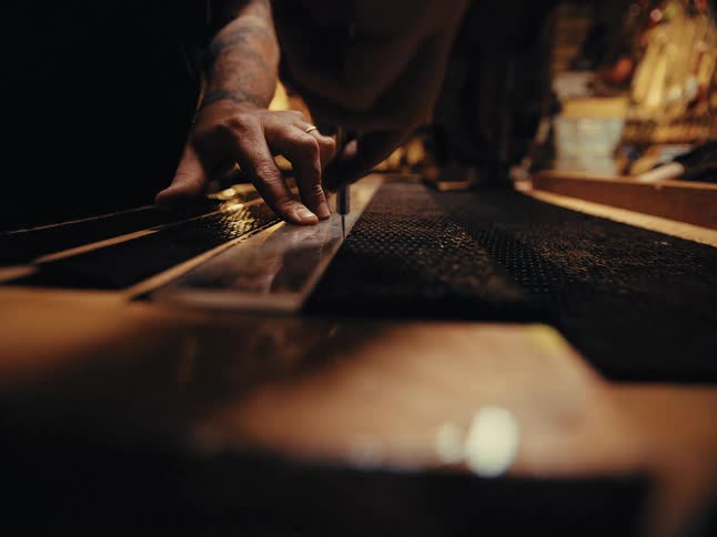 A craftsman with tattooed arms carefully cuts a piece of black textured material with a blade and ruler in a dimly lit workshop