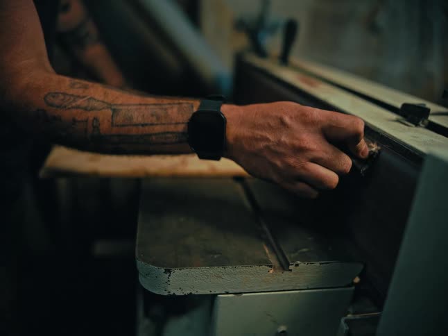 Close-up of a tattooed woodworker's hands using an industrial belt sander to smooth a wooden board in a dimly lit workshop