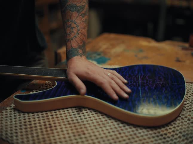 A luthier with tattooed arms carefully fits the neck to the body of a custom-built blue flame-top electric guitar
