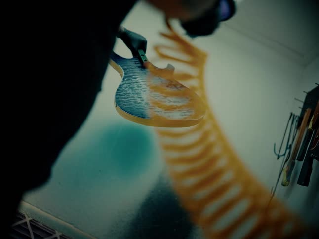 Low-angle seamless loop of a luthier spray painting a custom electric guitar body in a moody workshop