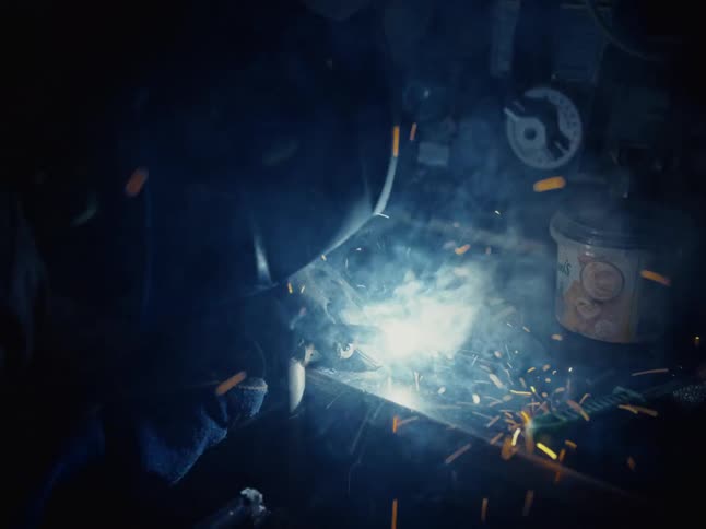 A welder in a protective mask works with metal, creating a cascade of sparks and smoke in slow motion