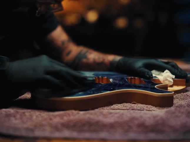 A craftsman with tattoos wearing black gloves meticulously polishes the custom blue body of an electric guitar in a workshop
