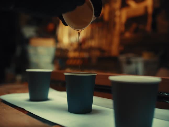 A craftsman in black gloves carefully pours golden epoxy resin from one paper cup into another in a dimly lit workshop