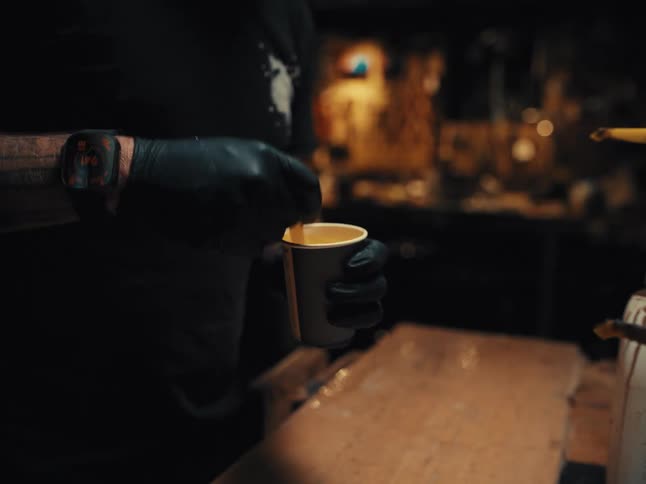 A craftsman wearing black gloves carefully mixes epoxy resin in a paper cup inside his workshop