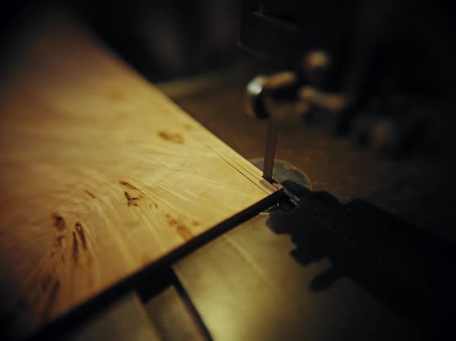 A bandsaw blade makes a precise cut along a marked line on a beautiful piece of wood in a dimly lit workshop