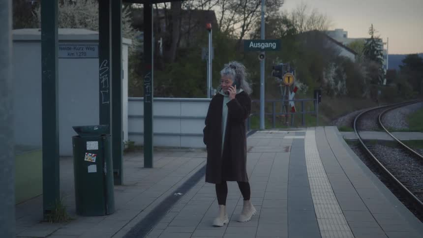 A woman with gray curly hair talks on her phone while waiting at a quiet train station in the evening