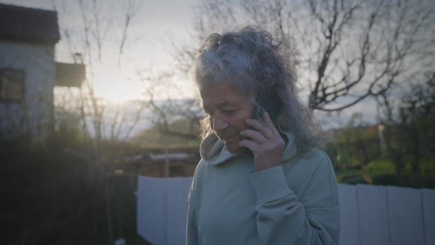 An elderly woman with gray hair stands in her garden at sunset, answering her smartphone with a concerned look