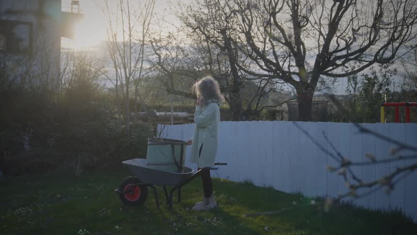 A mature woman takes a phone call while standing next to a wheelbarrow in her backyard during a golden sunset