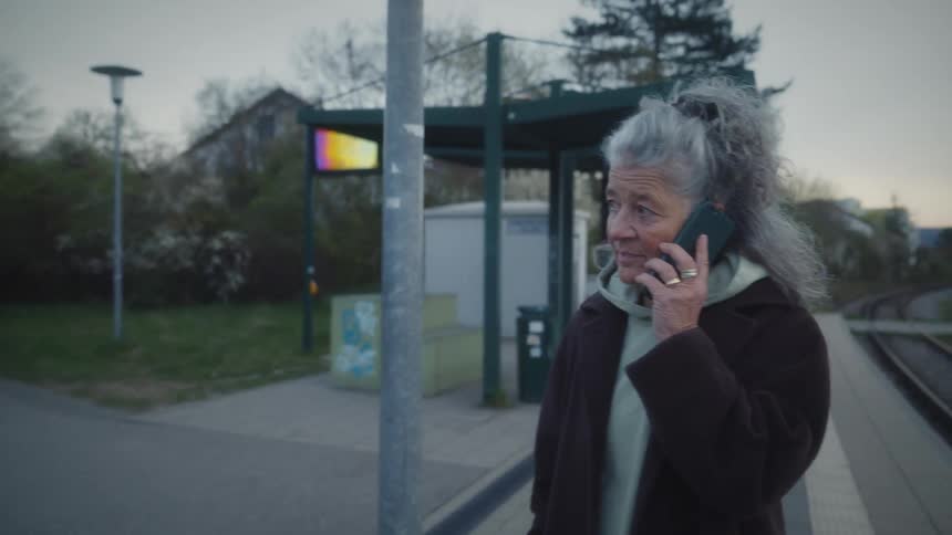 An elderly woman with gray hair talks on her smartphone while waiting at an empty train station platform at dusk