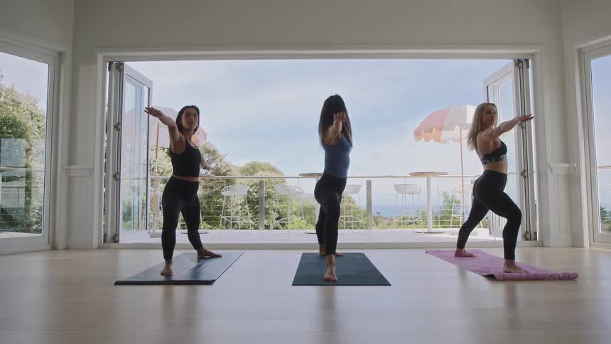 Three women practice yoga poses in unison in a bright studio with large open doors overlooking the ocean