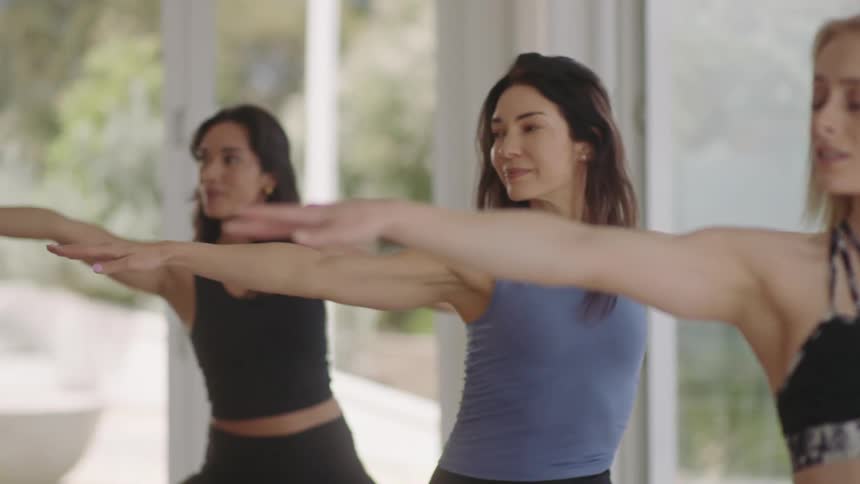 Three focused women in athletic wear hold the Warrior II yoga pose in a sunlit room