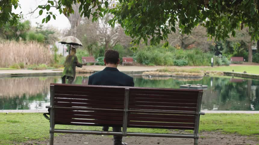 A man sits alone on a park bench, looking out over a still pond on a gloomy, overcast day