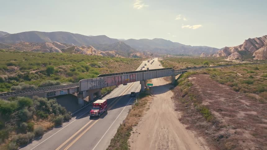 Aerial view of an ambulance driving along a rural highway in a desert landscape under a graffiti-covered railroad bridge