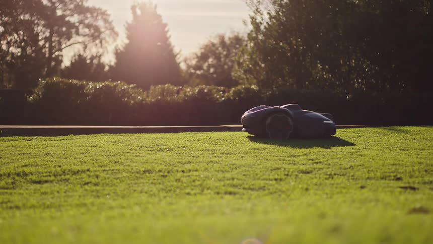 An automated robotic lawn mower trims a lush green yard as the golden hour sun shines in the background