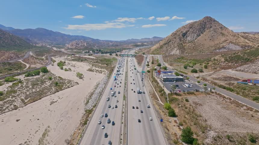 An aerial view of a busy multi-lane highway with heavy traffic flowing through a dry, mountainous desert landscape on a sunny day