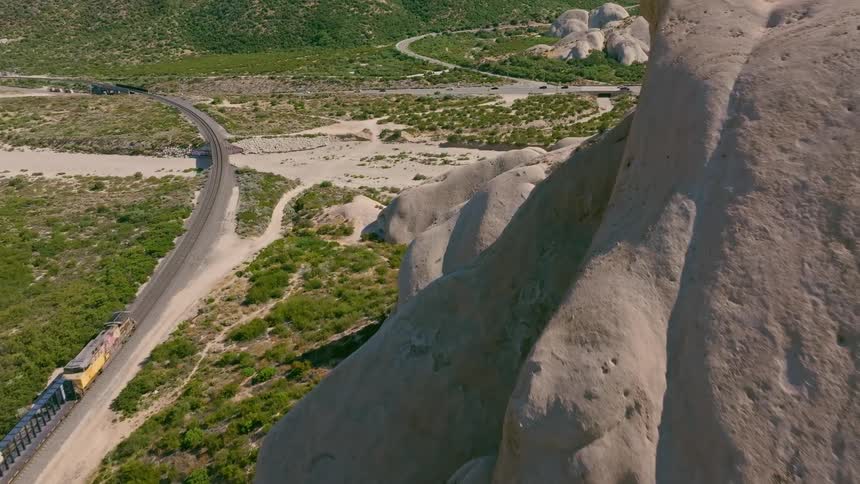 An aerial view shows a long freight train traveling on a railway alongside massive rock formations in a sunny desert landscape