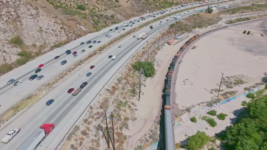An aerial view captures the parallel journeys of a long freight train and bustling highway traffic through a sunlit canyon