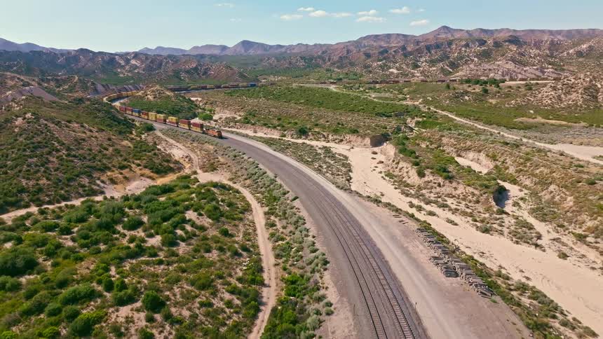 A long and colorful freight train travels along a winding track through a vast, arid mountain valley in this dynamic aerial shot