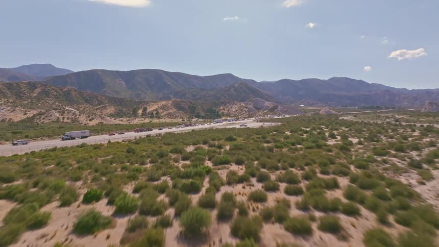 A fast-paced drone shot flies under a graffiti-covered bridge in a dry riverbed, soaring upwards to reveal a busy highway in the desert mountains
