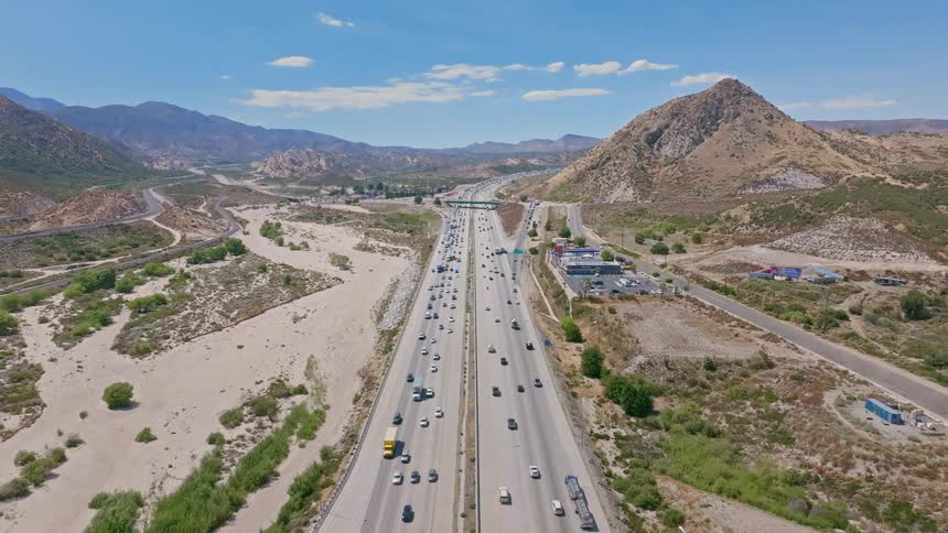 A static aerial view of busy traffic flowing along a multi-lane freeway that cuts through a dry, mountainous landscape