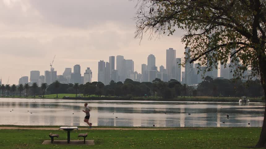 A man jogs along a scenic lakeside path in a city park with a hazy skyline in the distance