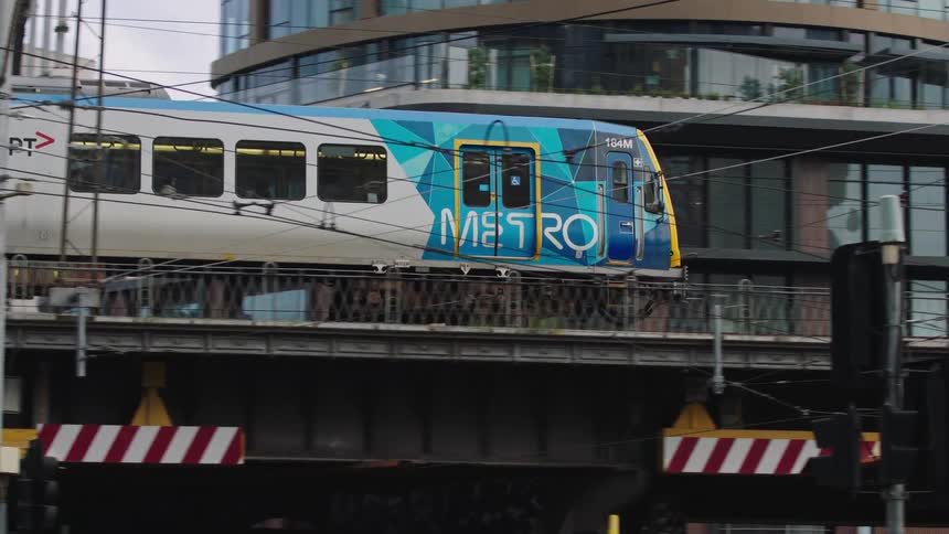 A slow-motion tracking shot follows a Metro train as it crosses an elevated bridge in an urban city setting