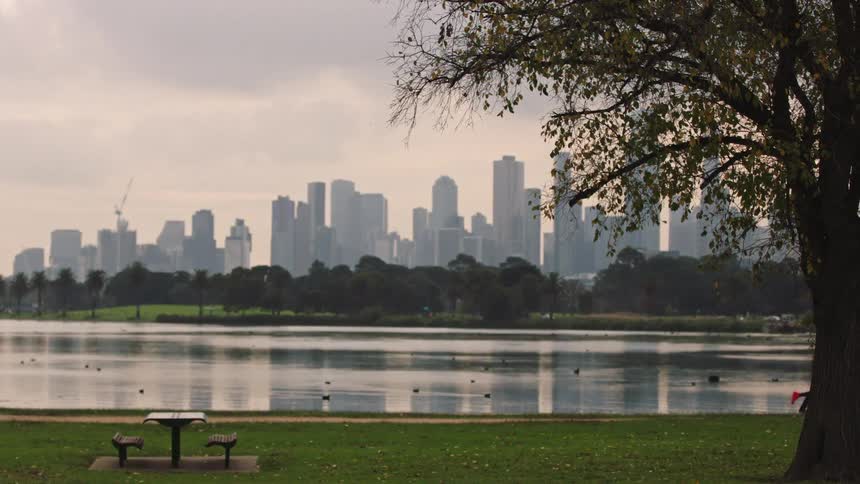 People exercise in slow motion in a park by a lake with a modern city skyline in the background on a cloudy day