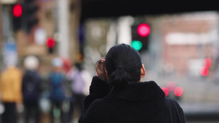 From behind, a woman in a black coat patiently waits for the traffic light to change on a busy city street