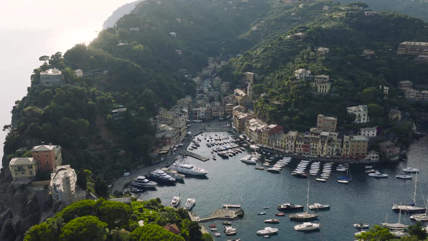 An aerial drone shot flies over the lush green hills towards the iconic harbor of Portofino, Italy, filled with luxury yachts and colorful buildings