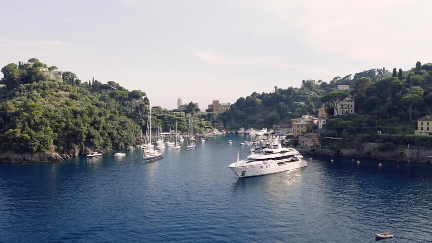 A wide shot of a luxury superyacht and other boats anchored in the tranquil bay of Portofino, Italy, surrounded by lush green hills