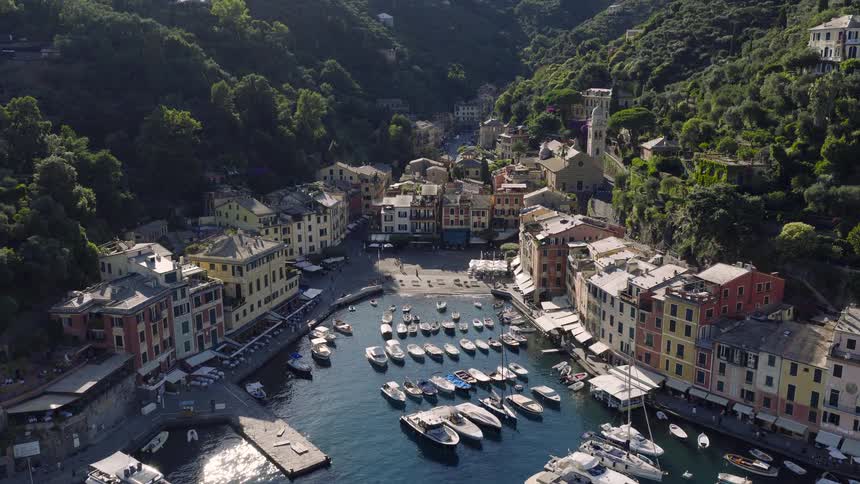 An aerial drone view slowly pushes into the famous and luxurious harbor of Portofino, Italy, showcasing the colorful buildings, docked yachts, and scenic coastline