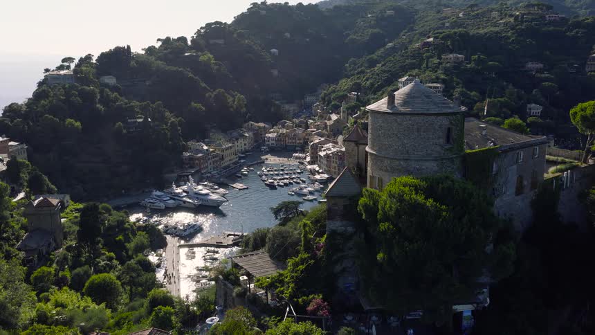 Aerial view reveals the iconic Portofino harbor with luxurious yachts, nestled below the historic Castello Brown on the Italian Riviera