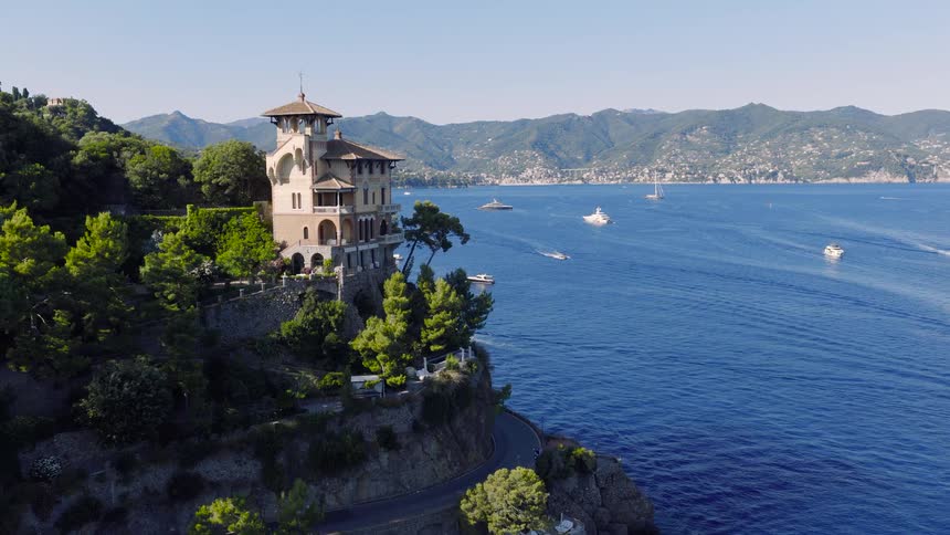 Stunning aerial shot slowly approaches a magnificent villa perched on a cliffside along the Italian Riviera, surrounded by luxury yachts on the sparkling blue sea