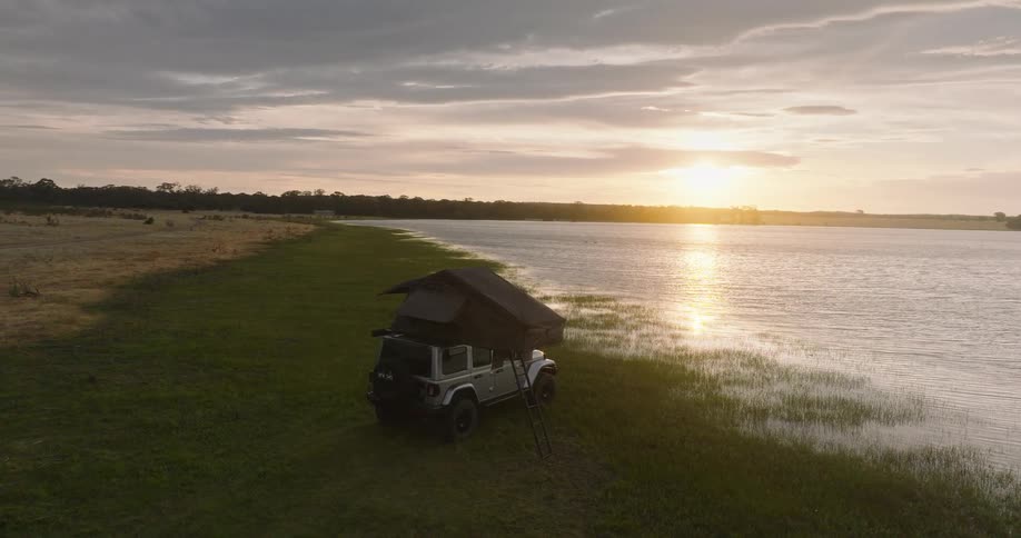 Aerial view of a 4x4 vehicle with a rooftop tent set up for camping on the grassy shore of a vast lake at sunset