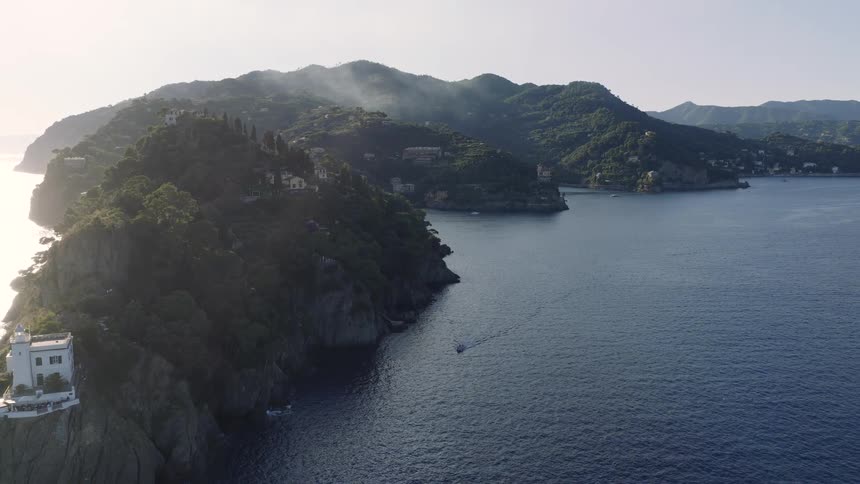 An aerial pull-back reveals the iconic Portofino Lighthouse perched on a rocky cliff overlooking the serene Ligurian Sea at sunrise