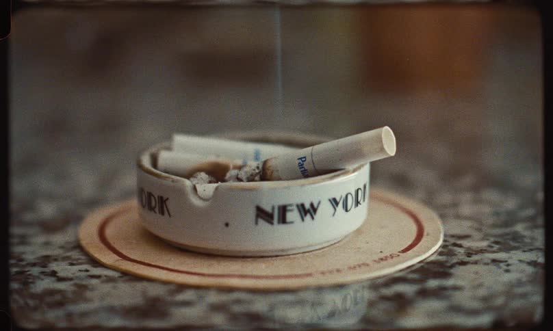 Smoke gently rises from a lit cigarette in a vintage New York ashtray in a grainy, retro-style close-up