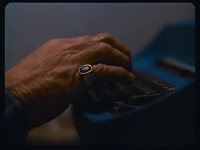 An intimate close-up shot on Super 8 film shows an elderly person's hands diligently typing on a vintage blue typewriter